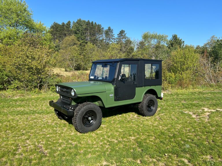 A ROXOR4 Full Hard Enclosure Cab Kit by Hardcabs is shown on grassy terrain, featuring a rugged, green boxy off-road look with black wheels and frame, surrounded by dense greenery under a clear blue sky.
