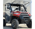 A 2018+ Polaris Ranger XP 1000 UTV with a Full Hard Cab Enclosure by Hard Cabs, featuring doors, front bumper, clear windshield, and roof frame, is parked outdoors near a light-colored building.