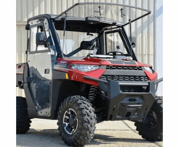A 2018+ Polaris Ranger XP 1000 UTV with a Full Hard Cab Enclosure by Hard Cabs, featuring doors, front bumper, clear windshield, and roof frame, is parked outdoors near a light-colored building.