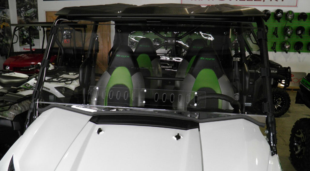 A Kawasaki Teryx 4 with a black roof and clear Lexan windshield (2016-20) is parked indoors. It features three black-and-green “Teryx” seats, and helmets are displayed on a green wall in the background.