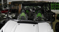 A Kawasaki Teryx 4 with a black roof and clear Lexan windshield (2016-20) is parked indoors. It features three black-and-green “Teryx” seats, and helmets are displayed on a green wall in the background.