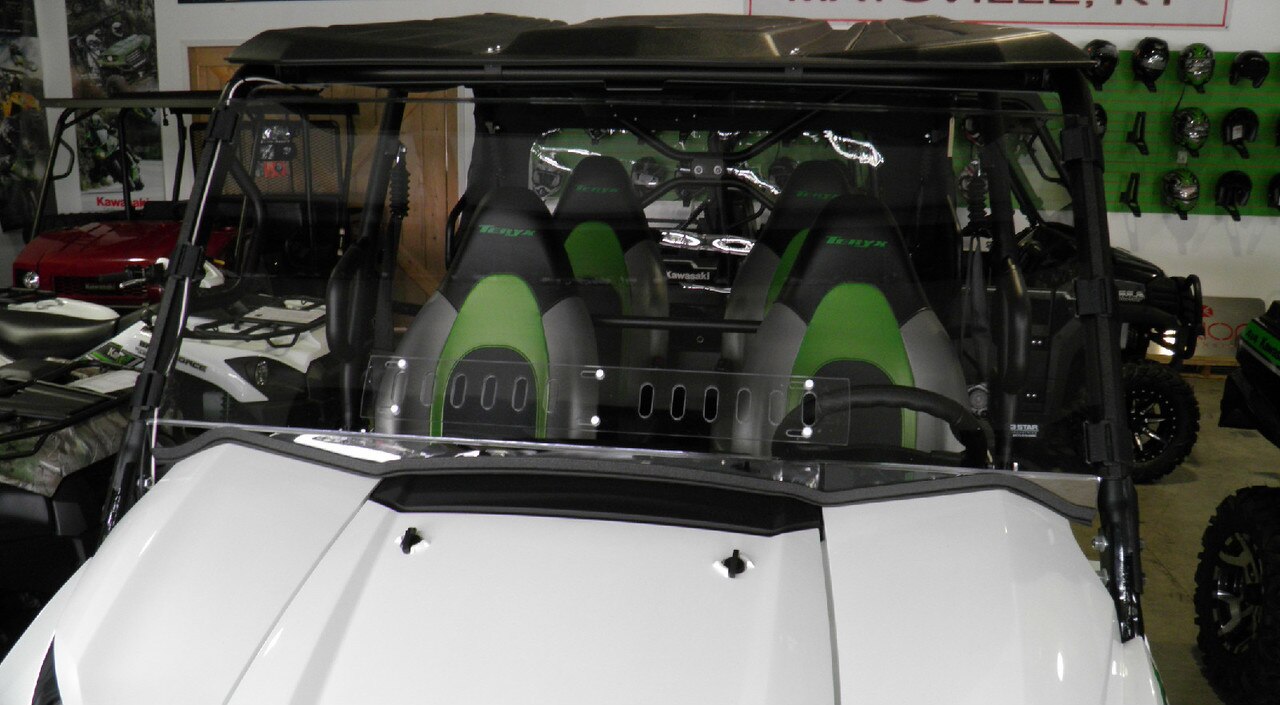 A Kawasaki Teryx 4 with a black roof and clear Lexan windshield (2016-20) is parked indoors. It features three black-and-green “Teryx” seats, and helmets are displayed on a green wall in the background.