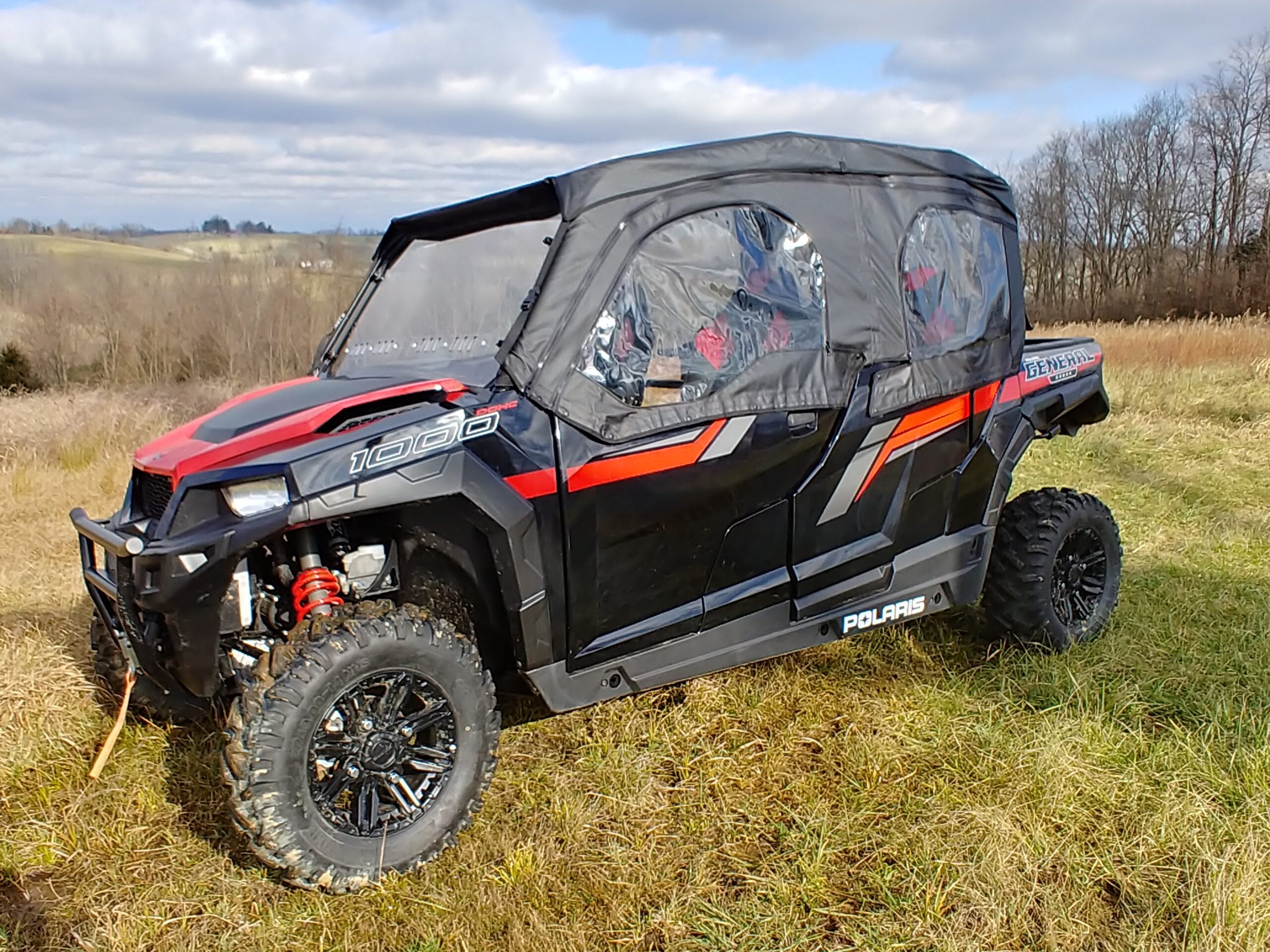 A Polaris General 4 with Offroad Armors Soft Doors Kit is parked on dry grass in a rural field. The side-by-side features large off-road tires, visible roll bars, and clear enclosures, with trees and cloudy skies in the background.