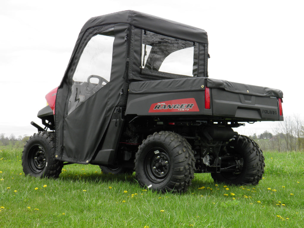 A Polaris Ranger with a Soft Full Cab Enclosure for an existing hard windshield is parked on grass, featuring clear windows and a cargo bed. The black utility vehicle is viewed from the rear passenger side on a cloudy day.