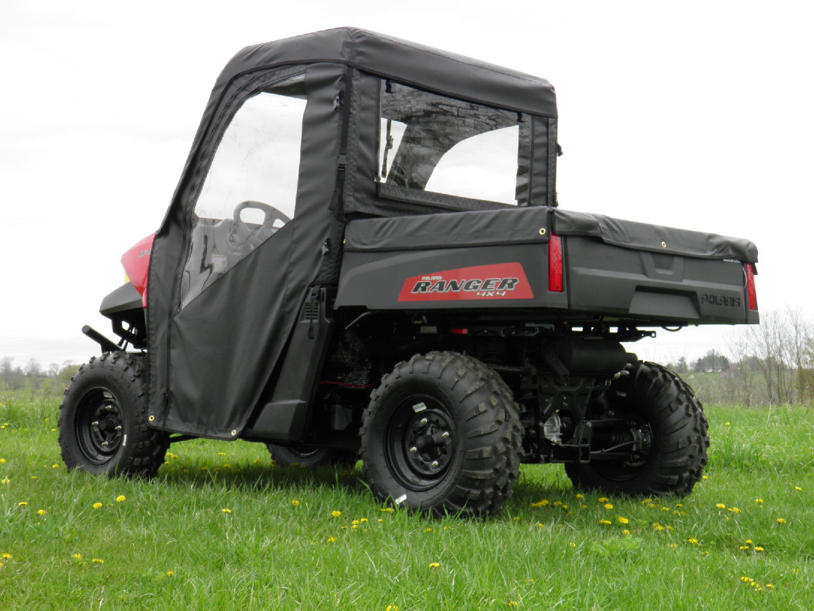 A Polaris Ranger with a Soft Full Cab Enclosure for an existing hard windshield is parked on grass, featuring clear windows and a cargo bed. The black utility vehicle is viewed from the rear passenger side on a cloudy day.
