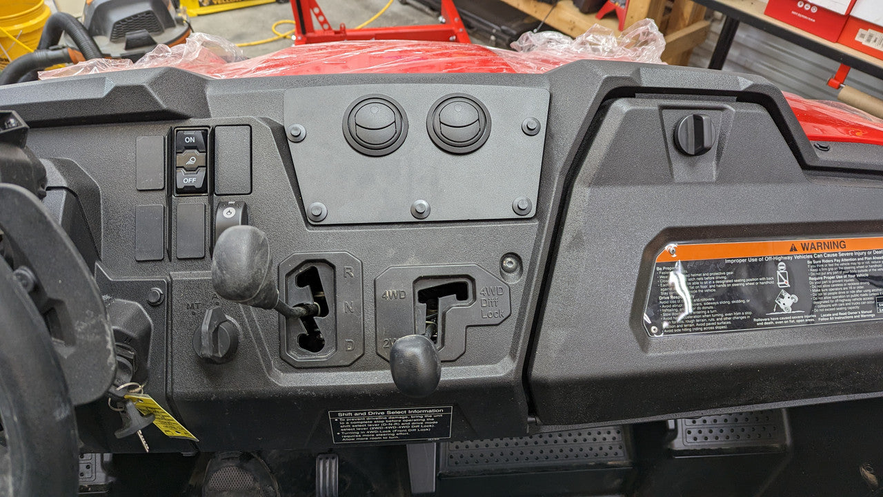 Close-up of a Honda Pioneer 700 (2023-2024) dash with the Ice Crusher Cab Heater installed, showing 2WD/4WD mode levers, switches, blank covers, a warning label on the right, and textured black plastic with some red body visible.