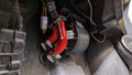 A close-up of a CFMoto Uforce 600 Cab Heater with Defrost (2021-2024) shows the cylindrical Ice Crusher Cab Heater with red hoses, metal clamps, and wiring in a car footwell; pedals and dust are visible.