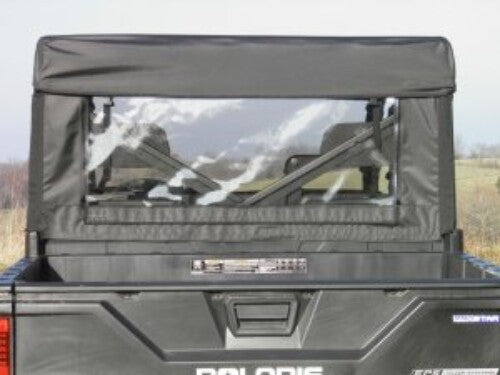 A Polaris Ranger Crew with a soft black fabric cab enclosure and a clear plastic rear window (Back Panel for all models). The empty cargo bed holds a small object near the cab. The vehicle sits on open grassy terrain beneath cloudy skies.