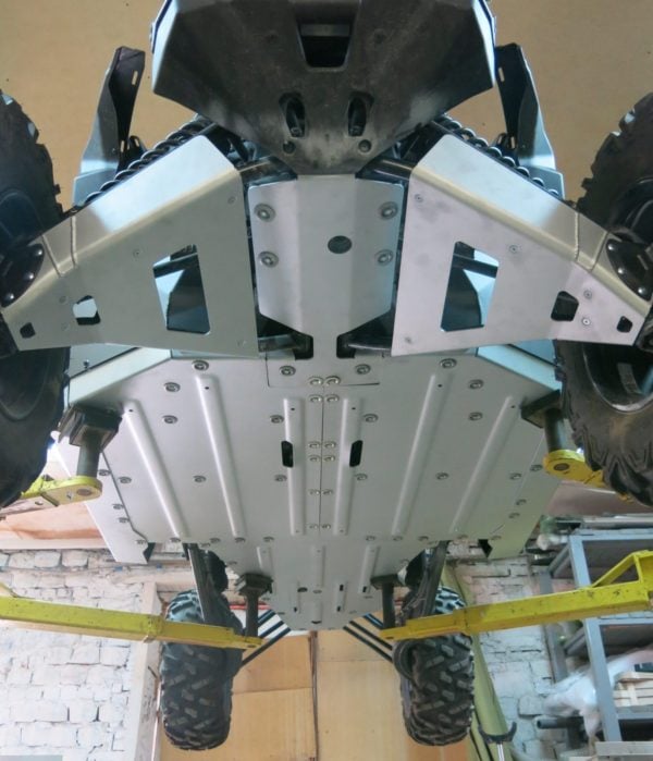 View from underneath a Can Am Maverick X3 64 / 72 Alloy Central Skid Plate on a hydraulic lift, displaying the full aluminum skid plate protecting the underside. Large tires and suspension arms are visible; workshop has brick walls and wooden doors.