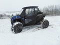 A Polaris RZR XP Turbo S with a Velocity Full Cab Enclosure for an existing hard windshield, in black and blue, is parked on snowy terrain with snow-covered tires, against leafless trees and wintry hills under a cloudy sky.