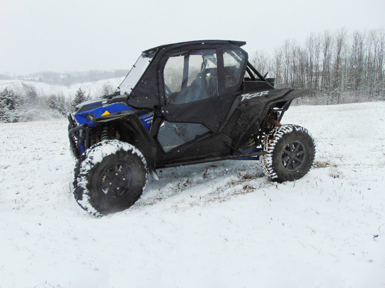 A Polaris RZR XP Turbo S with a Velocity Full Cab Enclosure for an existing hard windshield, in black and blue, is parked on snowy terrain with snow-covered tires, against leafless trees and wintry hills under a cloudy sky.