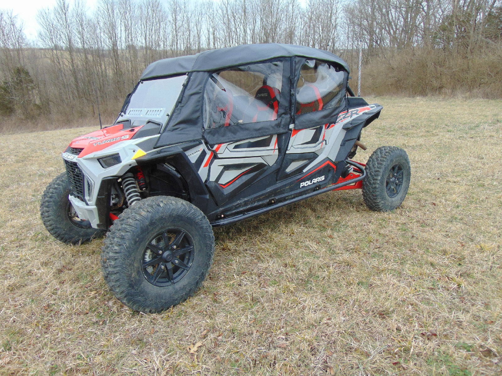 A Polaris Rzr Xp 4 Turbo S with a full cab enclosure for an existing hard windshield, featuring a black and red design, rugged tires, and red suspension components, is parked on dry grass under an overcast sky near leafless trees.