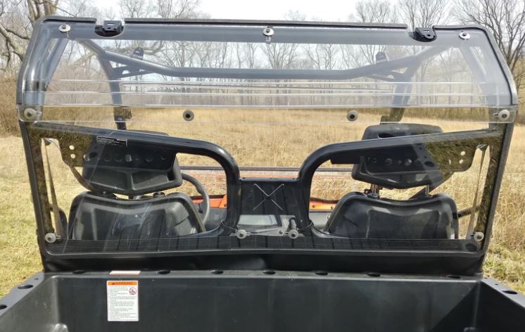 View from the cargo bed of a CFMoto UForce 500/800 equipped with a 2-piece Lexan Back Panel (includes soft lower panel with clamp and vent options), looking through the rear windshield at two black bucket seats; grassy field and overcast sky in the backgr