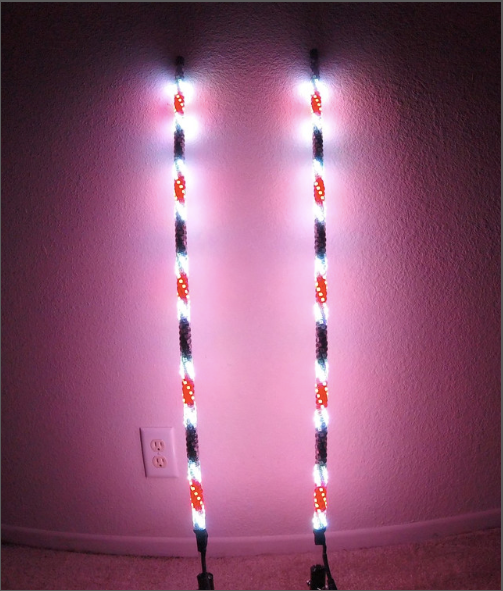 A Cyclone Whip - LVL 3 - Single with alternating red, white, and blue LED lights stands against a pink-lit wall, casting a colorful glow. A white electrical outlet and beige carpet are visible at the base of the wall.
