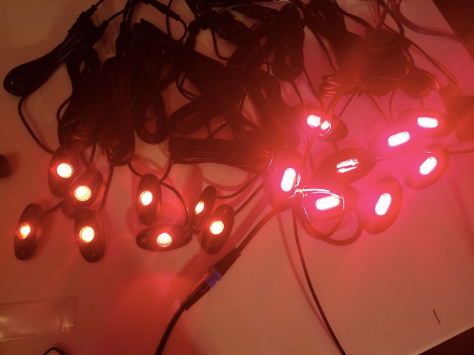 Several RGBW Rock Lights with black casings are arranged on a light surface, their LEDs glowing red and casting reflections, while black power cables lay tangled around them.