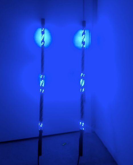 Two Cyclone Whip - Single LED light sticks emit vibrant blue light in a dim room, placed before a closed white door. Blue circles reflect on the door as a small green object rests on the gray floor between the glowing sticks.