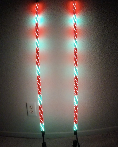 Two Cyclone Whips - 6FT- Pair stand side by side against a beige wall and carpeted floor, each emitting alternating red and green spiral lights. An electrical outlet is visible low on the wall to the left of the whips.