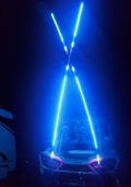 A vehicle is shown at night with two pairs of 6FT Woody Whips forming an X with blue LEDs at the rear, lighting up the scene. Part of another vehicle, likely an RV, appears on the left side of the image.