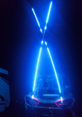 A vehicle is shown at night with two pairs of 6FT Woody Whips forming an X with blue LEDs at the rear, lighting up the scene. Part of another vehicle, likely an RV, appears on the left side of the image.