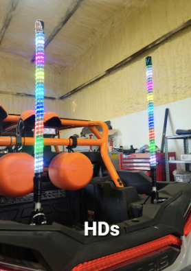 A utility vehicle is equipped with Cyclone Whips - 2FT - Pair, displaying multicolored LED patterns. Three orange round objects are on the back, HDs appears at the bottom center, and garage walls with tools and shelves are in the background.