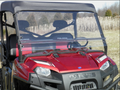 A Polaris Ranger Full Size 570 3-Passenger with a 2-piece clear windshield and black roof is parked on dry grass. The Polaris branding, grille, and headlights are visible, with tall grass and trees in the background under a clear sky.