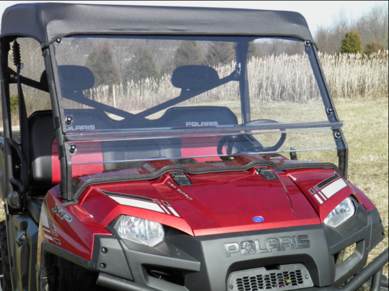 A Polaris Ranger Full Size 570 3-Passenger with a 2-piece clear windshield and black roof is parked on dry grass. The Polaris branding, grille, and headlights are visible, with tall grass and trees in the background under a clear sky.