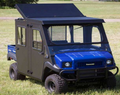 A 2013-2021 Kawasaki Mule Trans with an insulated steel top and raising rear panel, featuring a black cab, mesh windows, front grill guard, large off-road tires, and boxy shape, is parked on grass with trees in the background.