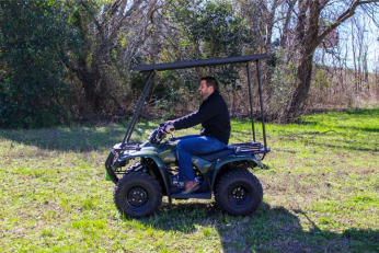 A man in a black jacket and jeans drives a green Honda Recon 250 Quad with the Steel Top Level 2 (Includes Headliner), which provides shade, across grassy terrain with trees and bushes in the background on a sunny day.