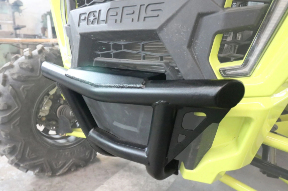Close-up of a Short Front Bumper Kit for Polaris RZR XP1000 (2019-2021) in yellow, showing the sturdy black metal brush guard with welded joints, triangular support design, and part of the rugged tire and grille in the background.