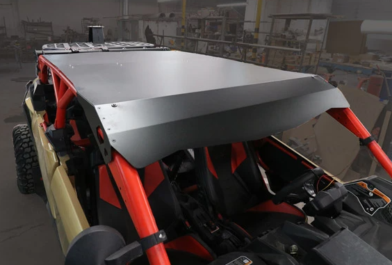A close-up of a rugged off-road vehicle featuring the Can Am X3 Max Roof and a red roll cage. Inside, two black-and-red bucket seats and the steering wheel are visible. The workshop background includes tools, shelves, and machinery.