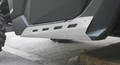 Close-up showing a Rock Sliders Kit for Can Am Maverick X3 2 Seater installed under an off-road vehicle. The silver guard features rectangular cutouts, is secured with bolts, and offers protection. Part of the tire and concrete ground appear in view.
