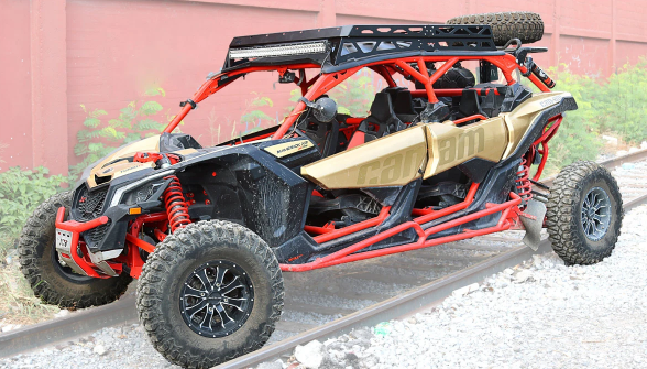 A gold and black Can-Am Maverick X3 Max 4-seater with a red frame, big all-terrain tires, visible suspension, and a Roof Rack Kit (with spare tire) is parked on railway tracks beside a red wall and green plants.