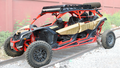 A gold and black Can-Am Maverick X3 Max 4-seater with a red frame, big all-terrain tires, visible suspension, and a Roof Rack Kit (with spare tire) is parked on railway tracks beside a red wall and green plants.