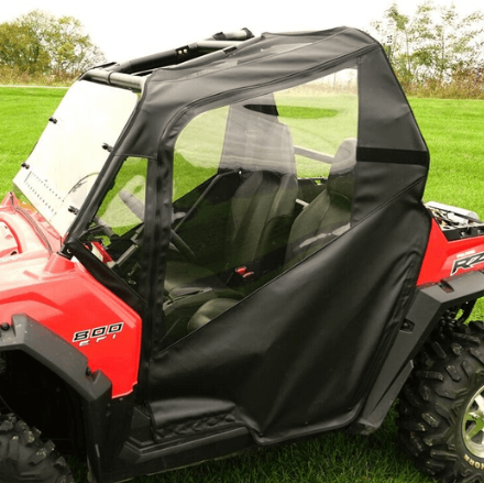 A Polaris Rzr 570/800/S 800/XP 900 fitted with Soft Full Doors featuring black fabric and clear plastic panels encloses two seats. The vehicle is parked on grass, with trees in the background.