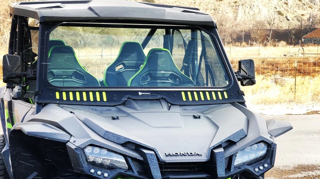 A rugged black Honda with green-accented seats and a Talon Vented Windshield With D.O.T Stamp is parked outdoors amid dry grass, bare trees, a fence, and rocky hills. The windshield features partial tinting and green vertical accents below.