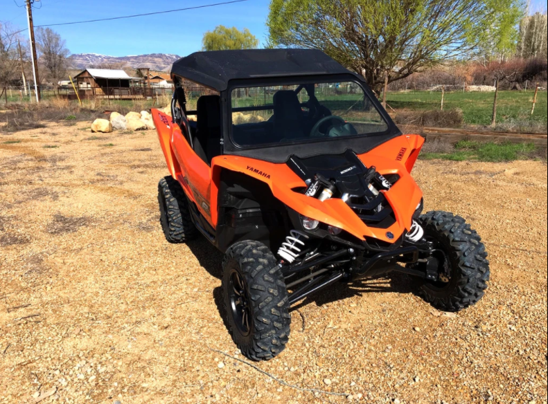 A Yamaha side-by-side with large tires, black roof, and a 2016-2017 Yamaha Yxz Full Front Glass Windshield With D.O.T Stamp is parked on gravel. Trees, fields, a fence, and buildings are visible under a blue sky.