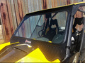 A Can Am Maverick Xrs with a D.O.T-stamped vented windshield is parked by a rusted metal wall. It features netted side windows and black-and-yellow bucket seats visible through the clear, certified front glass.