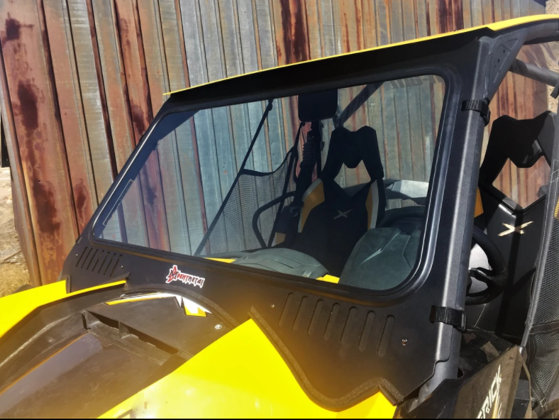 A Can Am Maverick Xrs with a D.O.T-stamped vented windshield is parked by a rusted metal wall. It features netted side windows and black-and-yellow bucket seats visible through the clear, certified front glass.