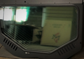 A large, tinted Replacement Glass with a D.O.T stamp leans against a wall, reflecting bright light and stacked cardboard boxes. The black frame features ventilation slits and a small Dynomax sticker.