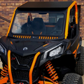 A Can-Am Mav-Trail/Sport/Commander with bold orange trim and rugged tires is parked by a brick wall, featuring the Vented Glass Windshield with D.O.T Stamp, visible suspension, roof, two seats, and distinctive safety bars.