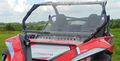 A close-up of a red off-road vehicle fitted with the Cf Moto Zforce 950 2-Piece Windshield, showing two empty seats against a green grassy landscape and cloudy sky, focuses on the vehicle’s front and top.