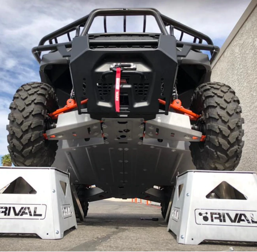 Low-angle view of a Polaris Ranger 1000 lifted on two white “Rival” branded ramps, showing the suspension and orange Alloy Central Skid Plate undercarriage against a cloudy sky and concrete wall.