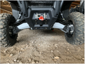 Close-up of Rival Powersports Can Am Defender HD5/HD8/HD10 Alloy Curved Front A Arm Guards installed under an off-road vehicle. The suspension, large tires, and metal guards are visible; photo taken in a sheltered area with a wooden ceiling.