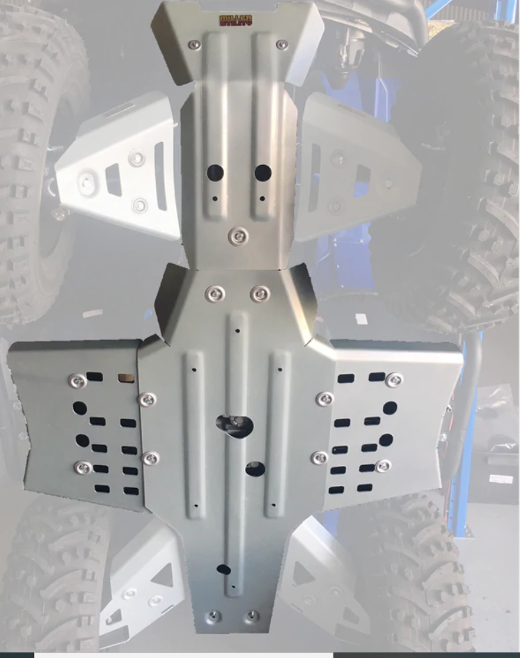 Close-up of a Yamaha Kodiak 450 Irs Alloy Central Skid Plate bolted under an off-road vehicle. The metal skid plate features ventilation holes and contoured edges, protecting the undercarriage with rugged tires visible in the background.