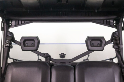 Close-up of the rear interior of a 2016+ CanAm Defender with black headrests, leather-style seats, support bar with bolts, and a Seizmik Hard Coated Rear Folding Windshield showing a hazy background through the rear window.
