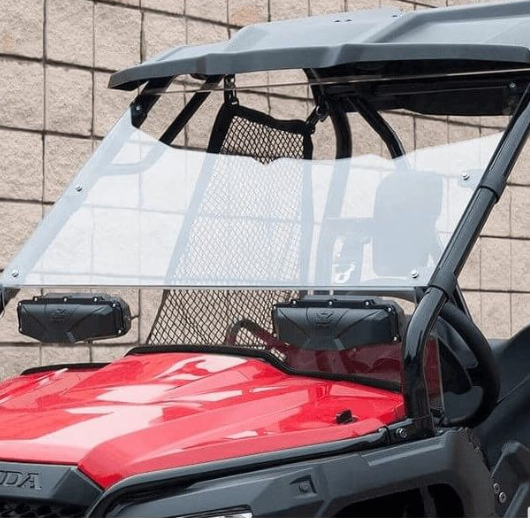 A close-up of a red Honda utility vehicle features the Seizmik Versa-Vent Hard Coated Front Windshield for Pioneer 500/700/1000, attached with black brackets, a mesh divider behind the seats, and a beige stone wall in the background. Focus is on the winds