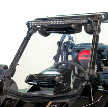 A close-up of a 2018+ Polaris RS1 Versa-Vent Hard Coated Front Windshield shows a black off-road vehicle with an LED light bar on top, plus a red-accented seat, steering wheel, and angular modern dashboard details inside.