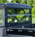 A close-up of the Seizmik Hard Rear Folding Windshield on a 2013+ Polaris Ranger 900/1000 Full Size Pro Fit shows its clear glass and sturdy panels, offering visibility to trees behind. The utility vehicle features a metal roll cage and angular lines.