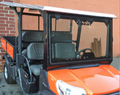 A Kubota RTV-X1140 with a diamond plated aluminum roof, black accents, clear windshield, two gray “Kubota” seats, mesh security grilles, and rear cargo bed is parked beside a brick and stucco wall.
