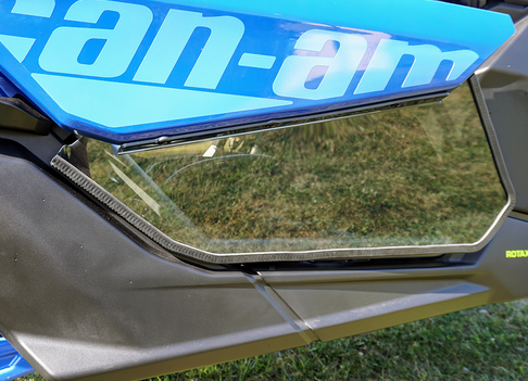 Close-up of Spike Lower Door Inserts Tinted on a Can Am Maverick X3. The blue door features tinted angular windows with a black and gray frame, reflecting grass and greenery in the background. Can-am is partially visible.